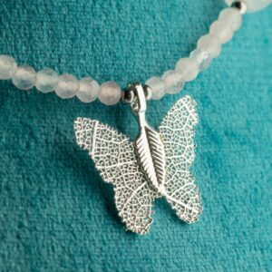 Collar de cuarzo rosa pálido en cuentas facetadas, con lascas lechosas y una mariposa metalizada sobre una hoja natural