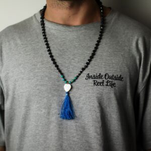 Collar largo de cuentas de piedra de lava con tramo turquesa, corazón blanco y borla azul, junto a su caja negra