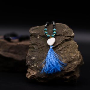 Collar largo de cuentas de piedra de lava con tramo turquesa, corazón blanco y borla azul, junto a su caja negra