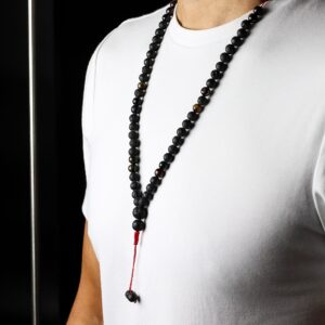 Collar largo de cuentas de piedra de lava negra y ojo de tigre con remate de cordón rojo y dos cuentas colgando, junto a su caja negra