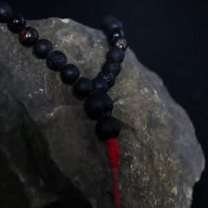 Collar largo de cuentas de piedra de lava negra y ojo de tigre con remate de cordón rojo y dos cuentas colgando, junto a su caja negra