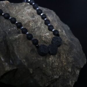 Collar largo de cuentas de piedra de lava negra con tres discos volcánicos en el centro y un tramo de cuentas beige y marrones, junto a su caja