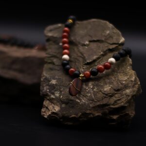 Collar largo de cuentas de lava negra y cuentas rojas con colgante ovalado de piedra rojiza envuelto en alambre dorado, junto a su caja