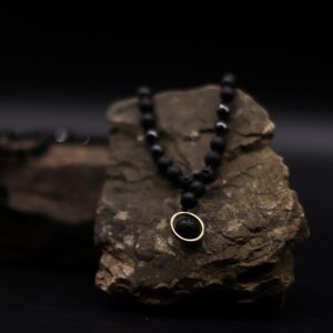 Collar negro de cuentas de piedra de lava y cuentas facetadas con colgante de una cuenta de lava dentro de un aro dorado, junto a su caja