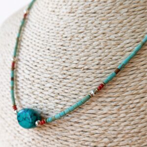 Collar fino de tubitos color turquesa verdoso con una piedra central ovalada de vetas negras y pequeños detalles rojos, plateados y dorados, sobre fondo blanco