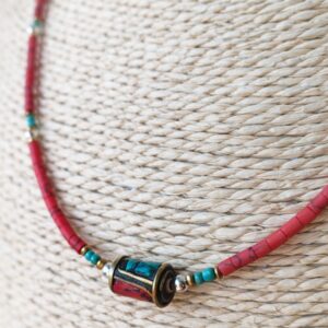Collar fino de cuentas rojo coral con toques turquesa y una cuenta central dorada con incrustaciones, puesto en un busto de rafia