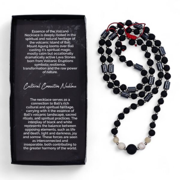 Collar largo de cuentas de piedra de lava negra y tubos grises con howlita blanca en el centro y nudo de macramé rojo, junto a su caja