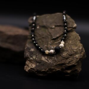Collar largo de cuentas de piedra de lava negra y tubos grises con howlita blanca en el centro y nudo de macramé rojo, junto a su caja