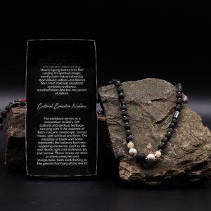 Collar largo de cuentas de piedra de lava negra y tubos grises con howlita blanca en el centro y nudo de macramé rojo, junto a su caja