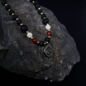 Collar de cuentas de piedra de lava con separadores dorados, cuentas turquesa y rojas y medalla redonda con el símbolo Om, junto a su caja