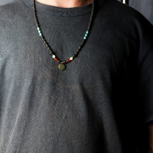 Collar de cuentas de piedra de lava con separadores dorados, cuentas turquesa y rojas y medalla redonda con el símbolo Om, junto a su caja