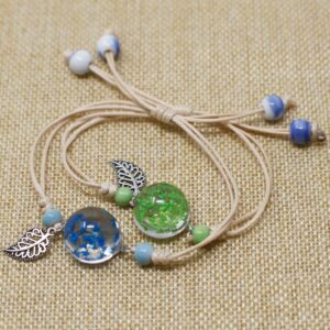 Dos pulseras de cordón beige con una esfera de vidrio con flores secas azules y verdes y una hoja calada plateada