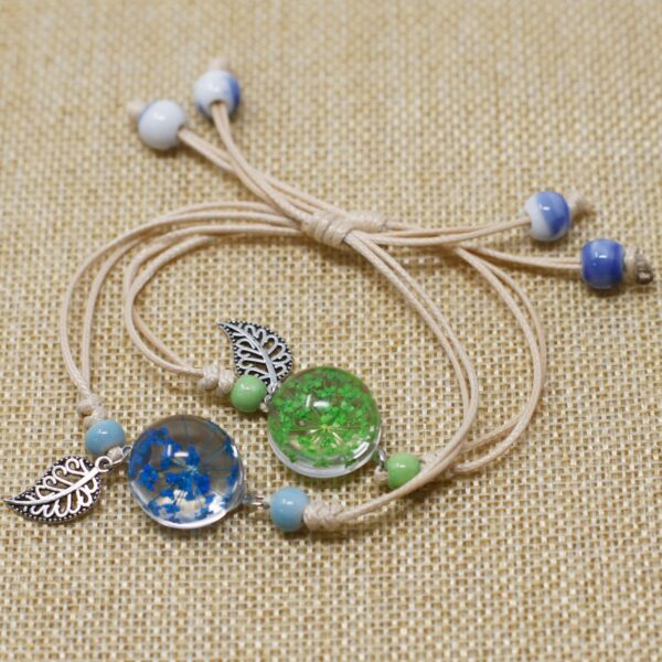Dos pulseras de cordón beige con una esfera de vidrio con flores secas azules y verdes y una hoja calada plateada