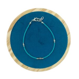 Pulsera fina de cuentas diminutas color turquesa con un tubito metálico grabado en el centro, mosquetón y cadenita, sobre fondo blanco