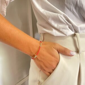 Pulsera fina de tubitos rojo coral con grupos de cuentas turquesa, bolitas doradas y una perlita blanca en el centro, sobre fondo blanco