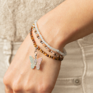 Set de tres pulseras elásticas en azul agua, melocotón y marrón dorado; la de ojo de tigre lleva una mariposa de tono plateado