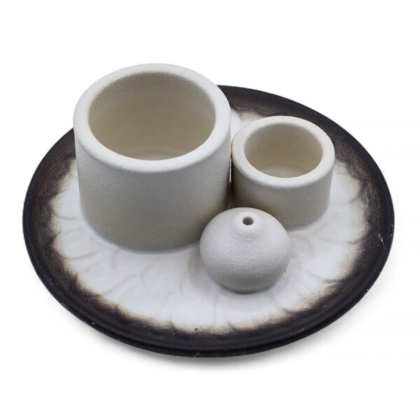 Soporte de cerámica blanco con dos cilindros y una bola portaincienso sobre un plato con relieve de pétalos y borde gris oscuro