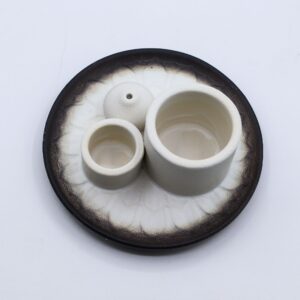 Soporte de cerámica blanco con dos cilindros y una bola portaincienso sobre un plato con relieve de pétalos y borde gris oscuro
