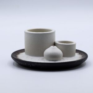 Soporte de cerámica blanco con dos cilindros y una bola portaincienso sobre un plato con relieve de pétalos y borde gris oscuro