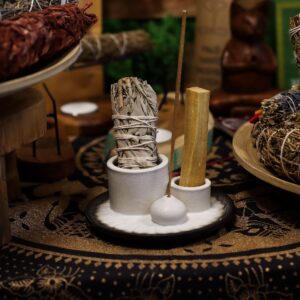Soporte de cerámica blanco con dos cilindros y una bola portaincienso sobre un plato con relieve de pétalos y borde gris oscuro