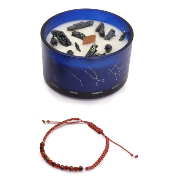 Vela de soja en vaso azul con constelaciones y trozos de pirita, junto a una pulsera de cordón rojo teja con cuentas de ojo de tigre y cornalina