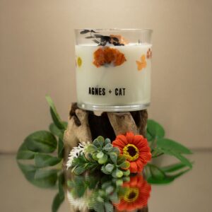 Vela de soja en vaso de vidrio con flores secas naranjas y hojas negras junto a su caja blanca