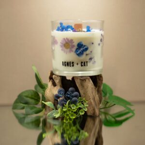 Vela de soja en vaso de vidrio con flores secas azules y lilas junto a su caja blanca