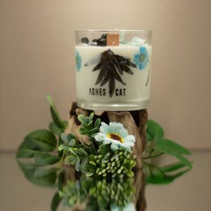 Vela de soja en vaso de vidrio con una hoja de helecho oscura y flores secas azul verdoso junto a su caja blanca