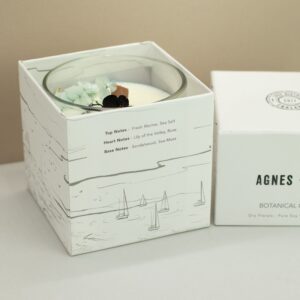 Vela de soja en vaso de vidrio con una hoja de helecho oscura y flores secas azul verdoso junto a su caja blanca